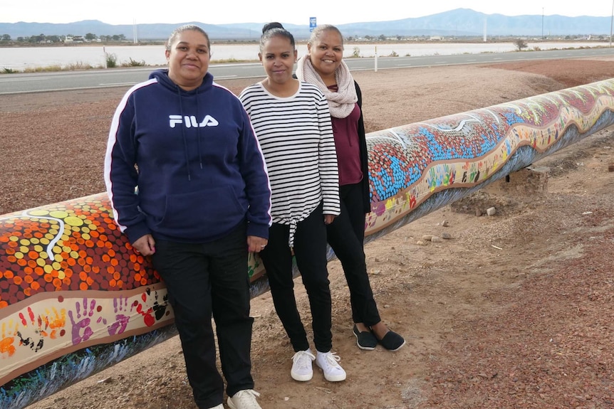 Three Aboriginal women stand in front of a pipeline next to a highway, that is painted.