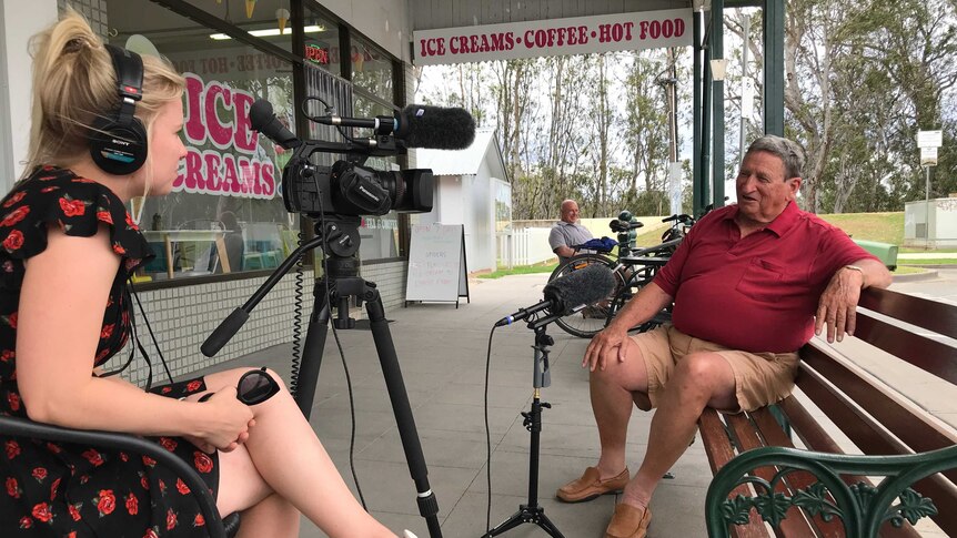 A young woman sits behind a TV camera interviewing an older man who sits on a park bench outside an ice cream shop.
