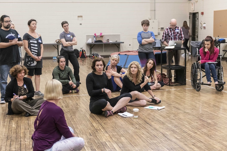 A group of people in a hall - all looking at a woman in the centre who is demonstrating a movement.