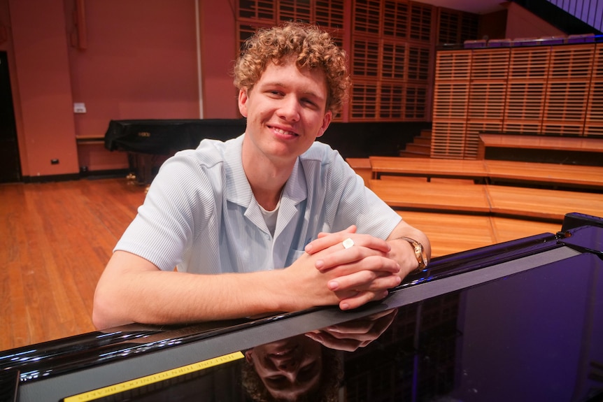 A young man smiles with his hands together resting on a piano.