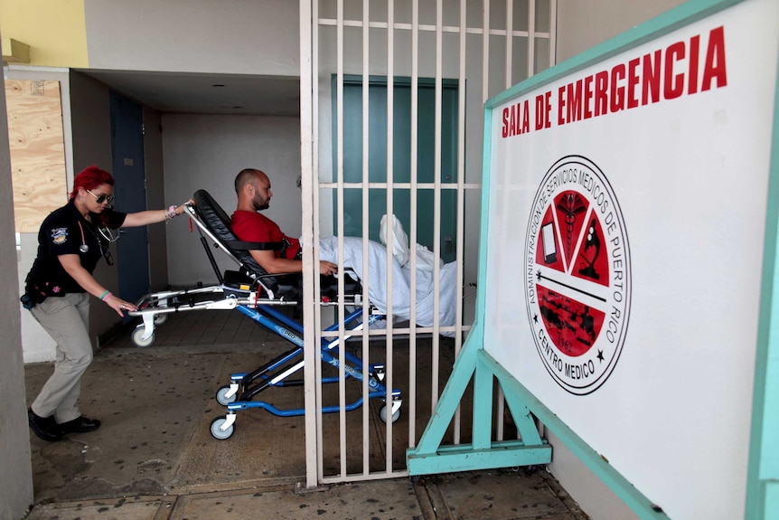 A paramedic pushes a man on a stretcher with a blanket over his knees past a gate into a medical centre.
