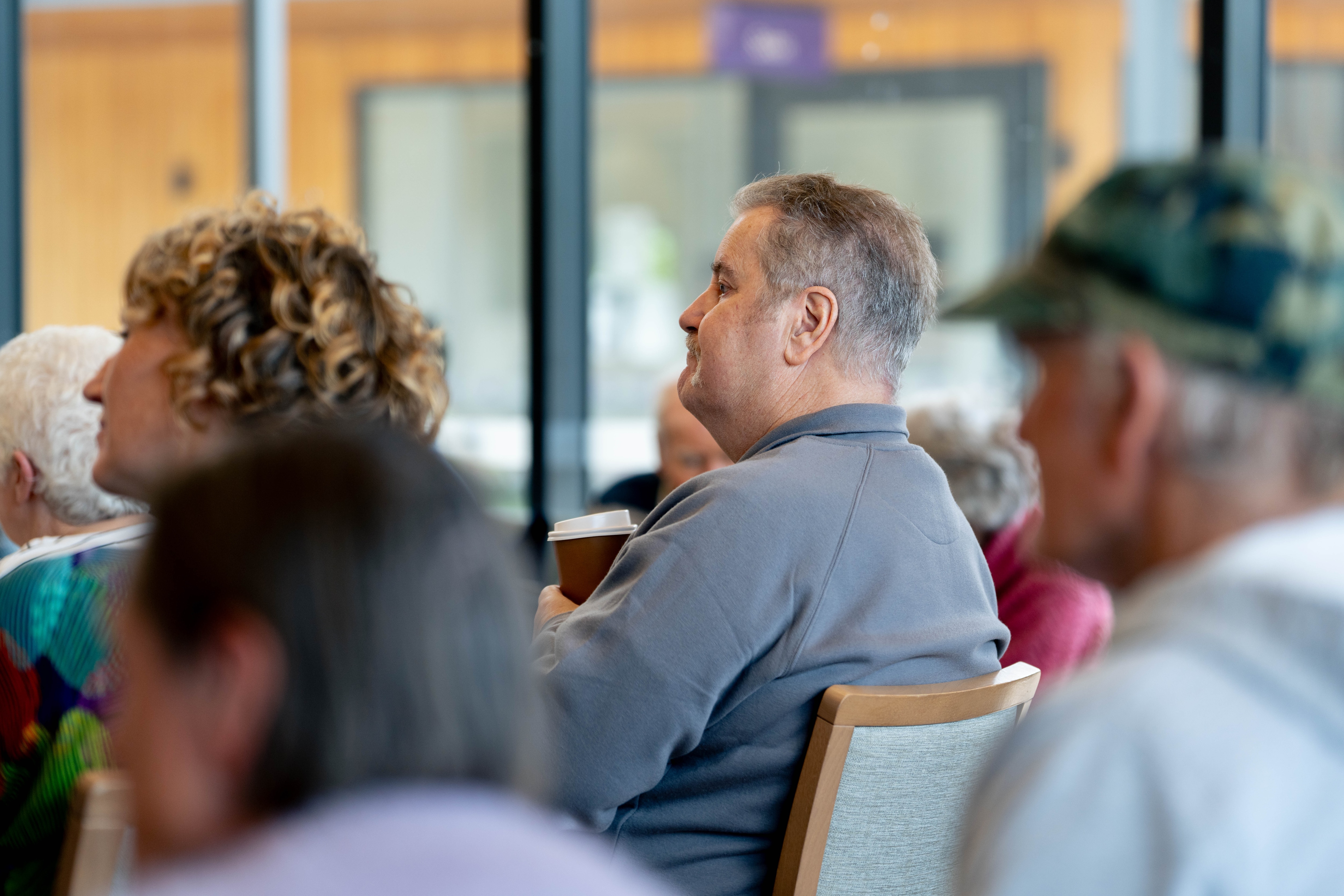 Mick, wearing a grey quarter zip jacket, sits in a row of chairs looking ahead, listening to music.