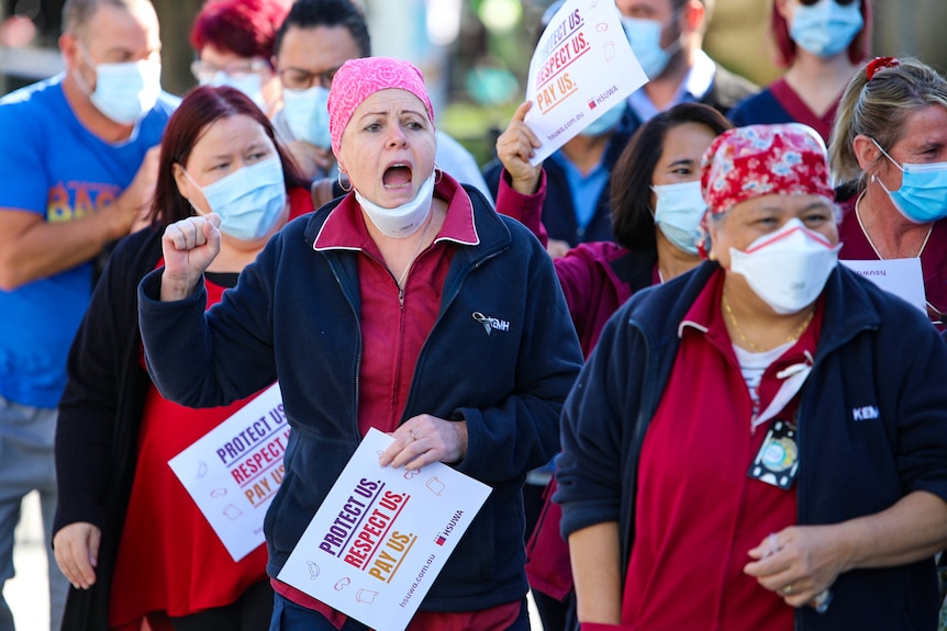 Тълпи от здравни работници в униформи и маски, държащи знаци.