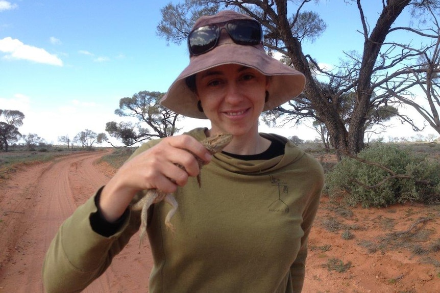 A woman in a long sleeve shirt and a bucket hat holds a small lizard in her fingers.