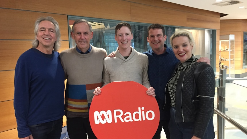 five people standing shoulder to shoulder, smiling for the camera, with the middle one holding a red round ABC radio sign