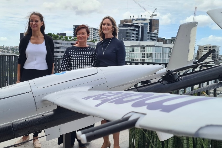 Three women stand next to a drone on a rooftop.