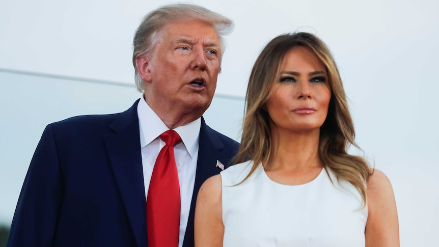 Donald Trump in a suit and red tie stands slightly behind melania trump in a white dress