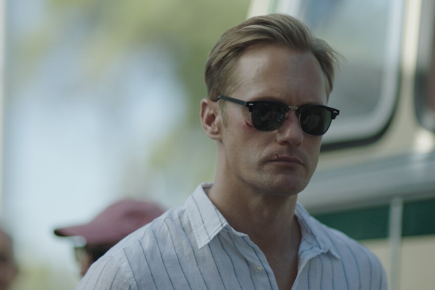 Alexander Skarsgård, a white man with blonde hair is wearing sunglasses and a white striped shirt. He has cuts on his face.