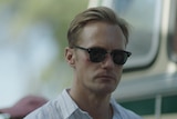 Alexander Skarsgård, a white man with blonde hair is wearing sunglasses and a white striped shirt. He has cuts on his face.