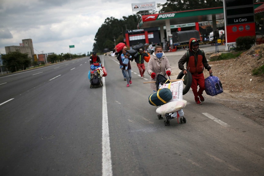 A group of people walk along the highway with luggage and face masks.