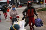 A group of people walk along the highway with luggage and face masks.