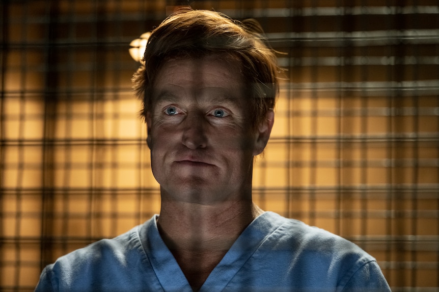 Man with short blonde hair and blue eyes wears blue prison garb and stands staring out behind metal bars.