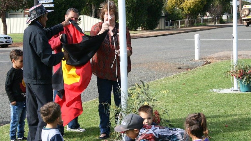 A group of people stand at a flagpole on a green lawn, raising the Aboriginal flag.