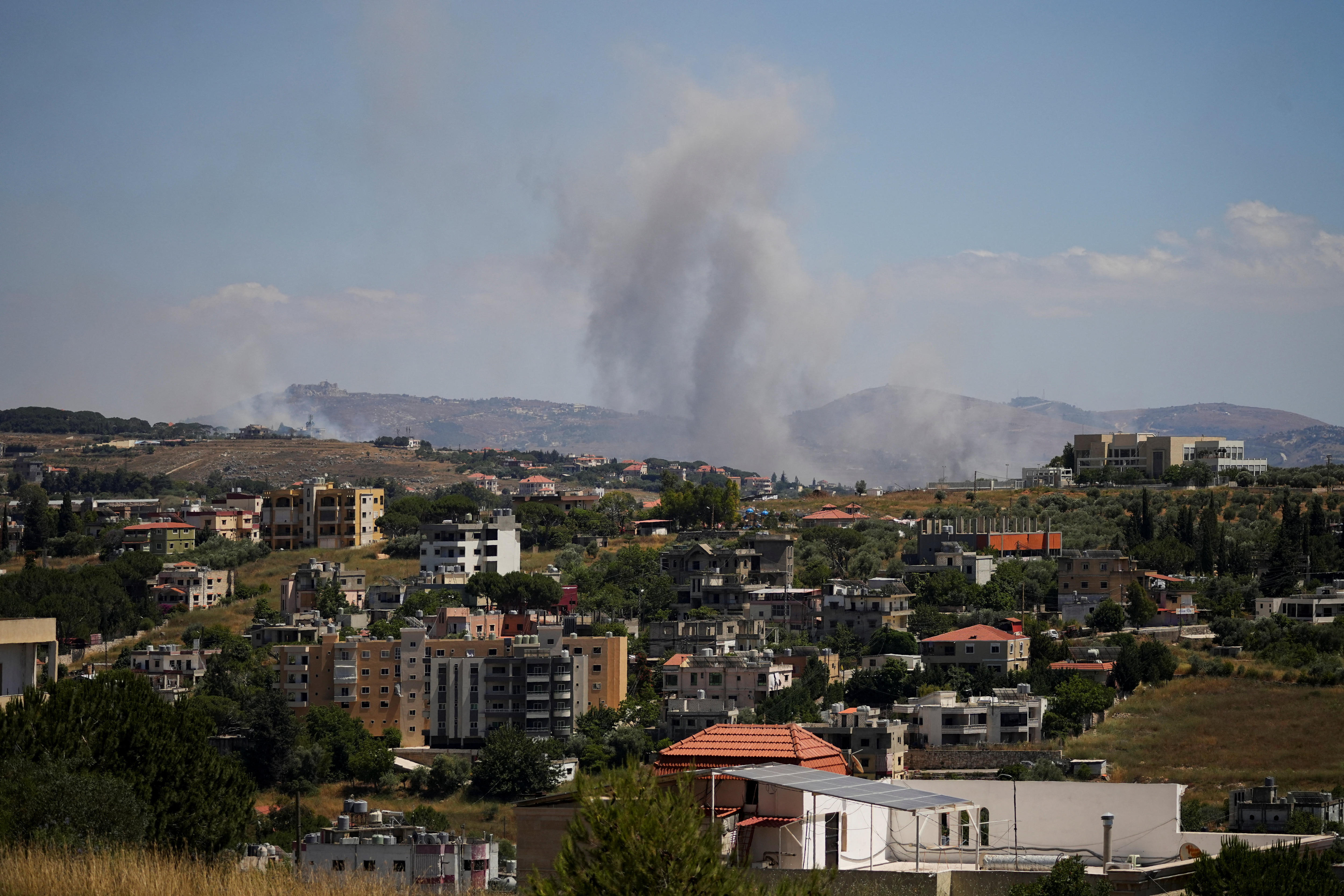Clouds of smoke can be seen over a hilly, green landscape with multiple multi-storey apartment buildings in it