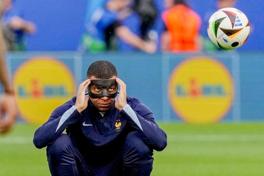 French football star Kylian Mbappé sits with his hands on a mask covering his face, as he watches a ball in the air. 