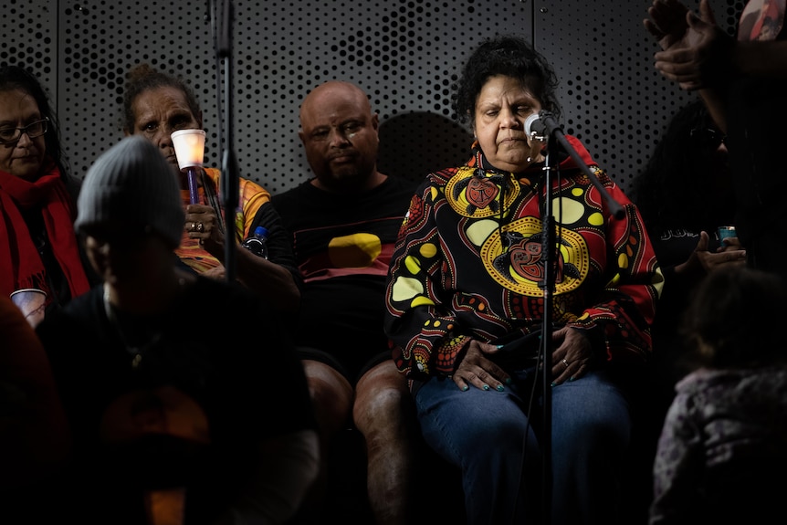 A woman sits down at a vigil, surrounded by people holding candles. 