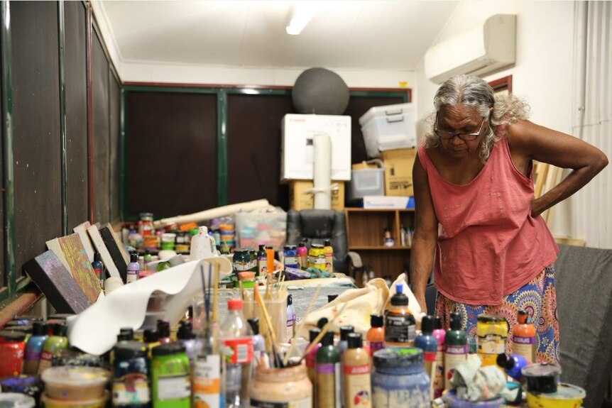 Senior indigenous woman stands looking down at her desk surrounded by bottles of paints, paintbrushes and canvas