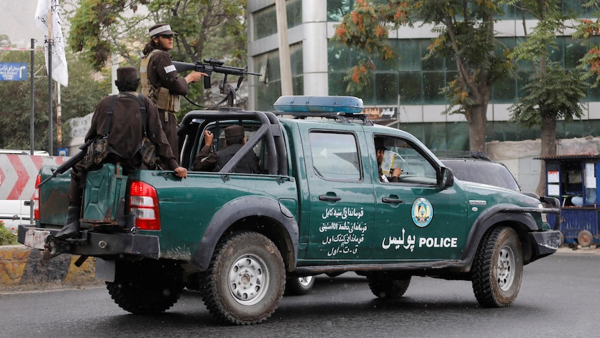 Two men stand in the tray of a green ute labelled POLICE. They are holding weapons and watching the street