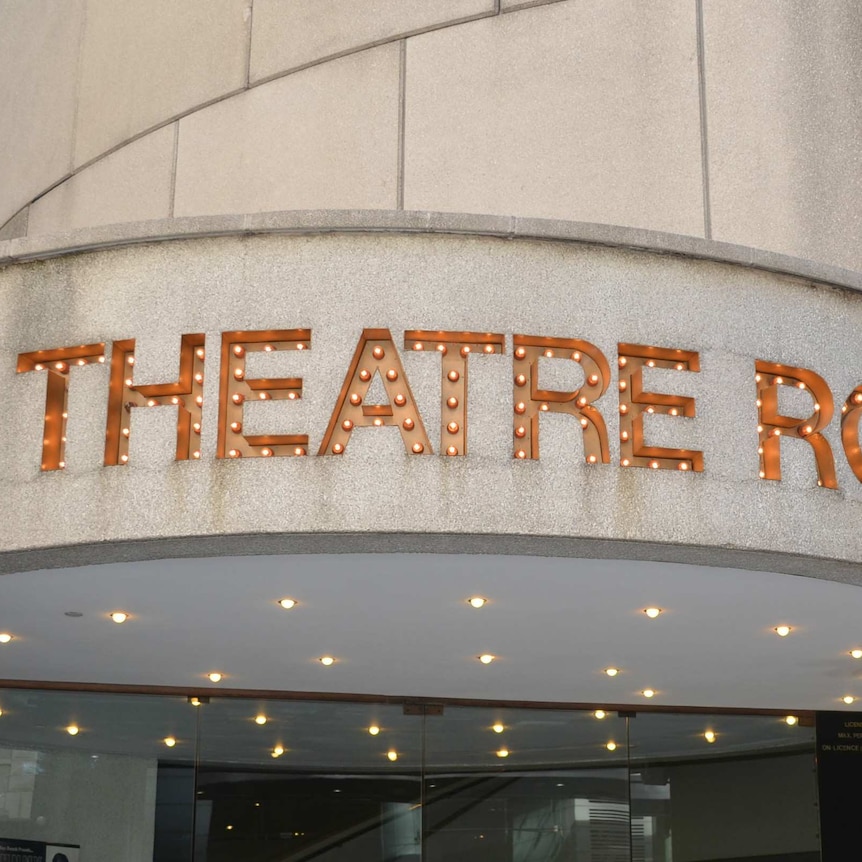 A sign made of lightbulbs that spells "THEATRE ROYAL" above a glass door.