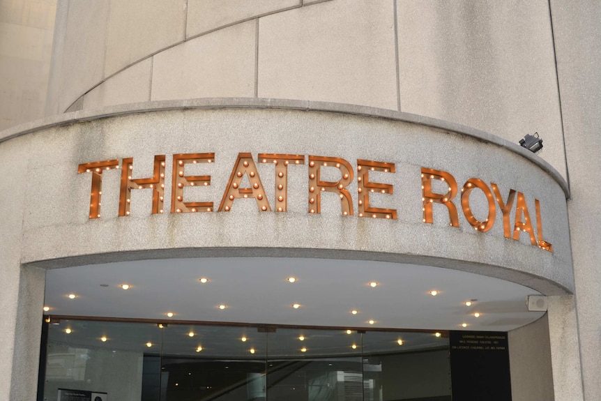 A sign made of lightbulbs that spells "THEATRE ROYAL" above a glass door.
