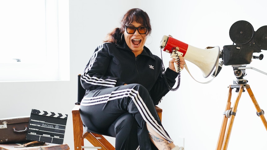 A woman in an black and white Adidas tracksuit sits on a director's chair holding a megaphone, surrounded by film props.