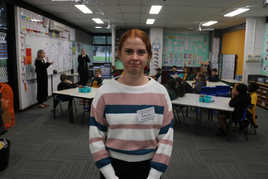 A woman standing in a classroom. She is wearing a striped jumper 