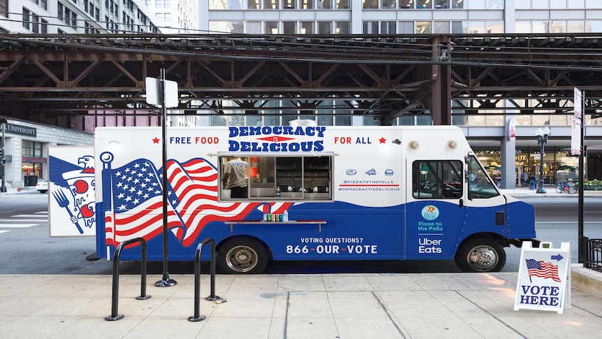 A red, white and blue themed food truck is parked on a NYC street, with a 'vote here' sign to the right.