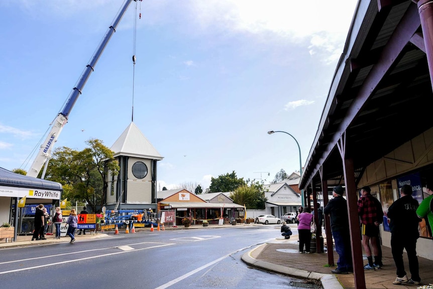 A crane lifting on the roof of a tower in the middle of a main street with many people looking on.