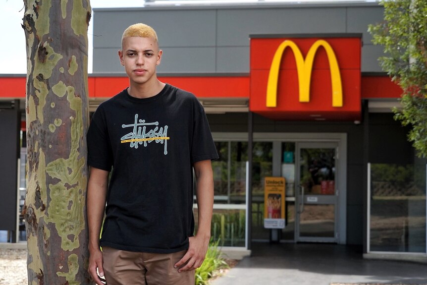 A young man with short blonde hair wearing a black tshirt leans against a tree. Behind him is a McDonald's