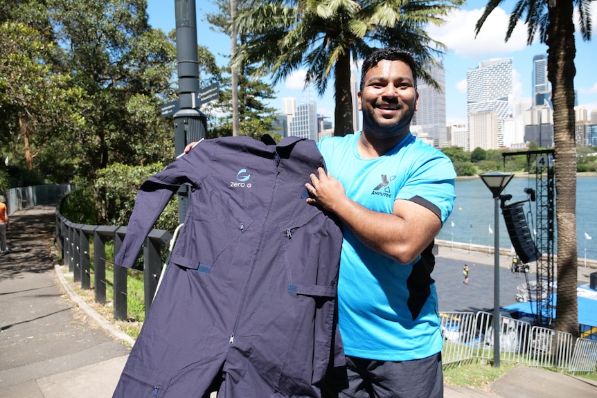 A man wearing a light blue t shirt smiles while holding a set of dark blue overalls