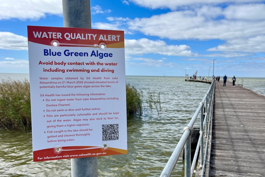 A sign on a pole in front of a boat ramp and jetty warning people to stay out of the water.