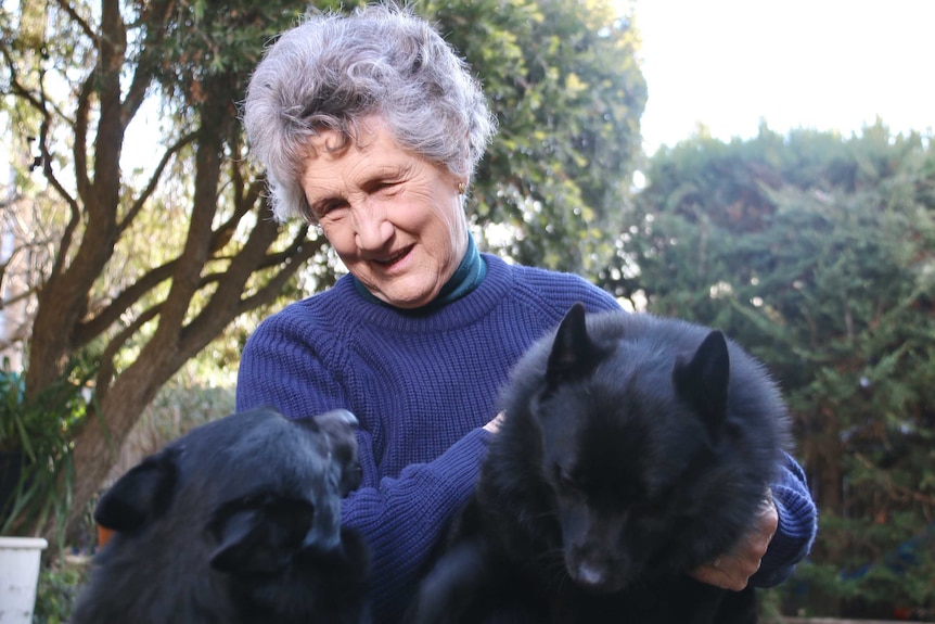 A woman with white hair and a purple jumper pats two black dogs in a garden.