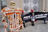 a skeleton made of prescription bottles and a skull stands next to people holding a sign protesting purdue and sackler family