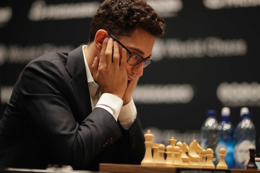 A young man wearing glasses and a black suit stares at chess board, resting his head on his hands.