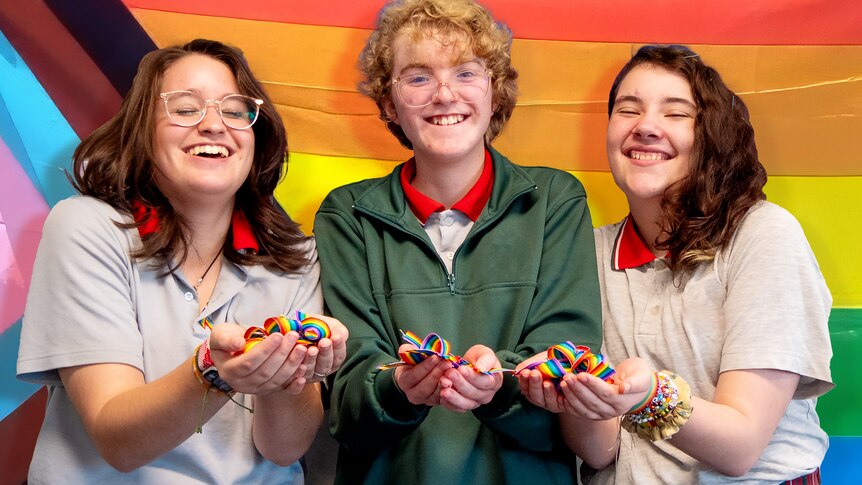 Three students smile and laugh while holding handfuls of rainbow ribbons. A pride flag in the background.