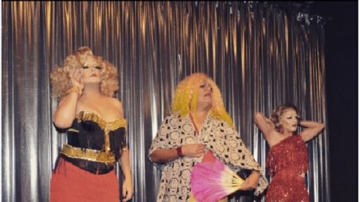 Three drag performers stand on a small stage with a silver curtain in the background.