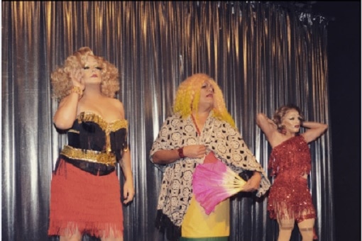 Three drag performers stand on a small stage with a silver curtain in the background.