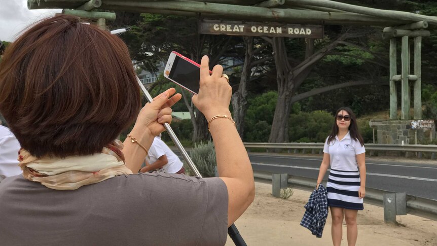 A woman poses for a photo under a sign that says "Great Ocean Road". In the foreground another woman takes her picture