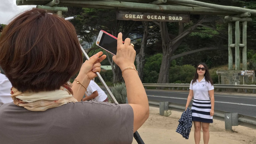 A woman poses for a photo under a sign that says "Great Ocean Road". In the foreground another woman takes her picture