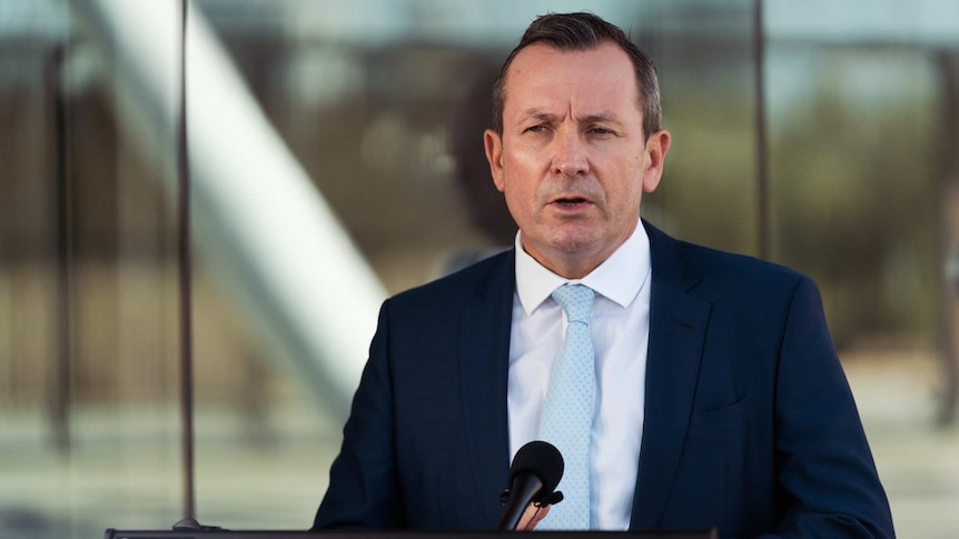 Mr McGowan speaks into a microphone in front of a podium, with reflective glass in the background.