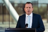 Mr McGowan speaks into a microphone in front of a podium, with reflective glass in the background.