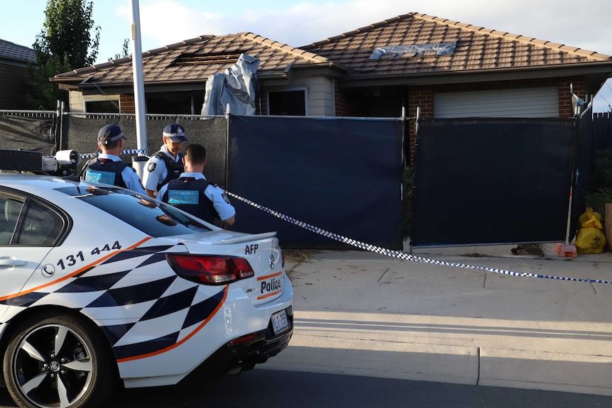 Police stand near their car while a house damaged by fire is obstructed by screens.