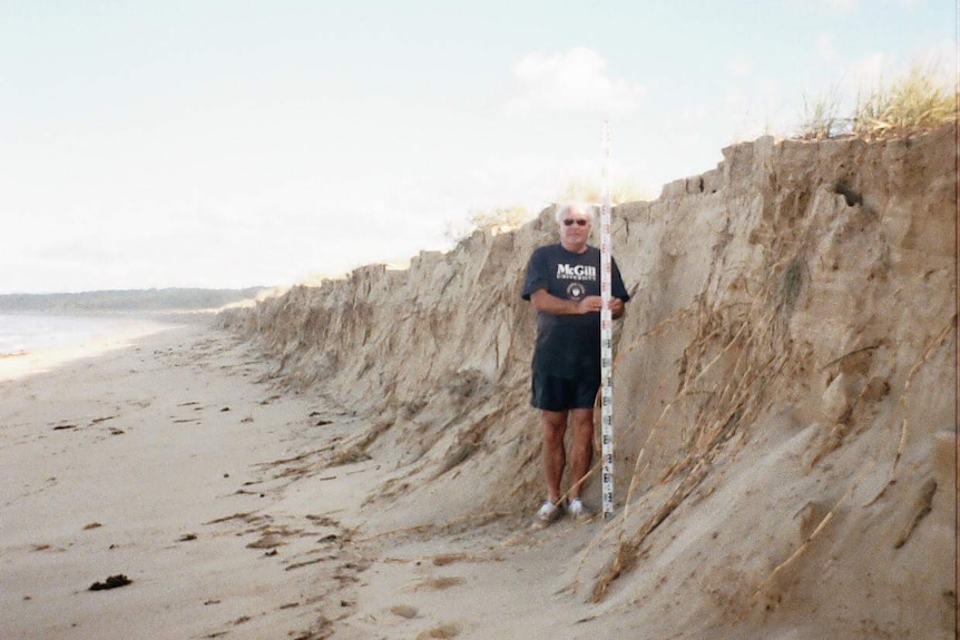 A man stands beside a sand dune cliff with a long white pole, measuring the height of the drop.