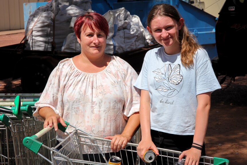 A woman with short red hair standing alongside a teenage girl in a blue t-shirt behind a shopping trolley filled with cans.