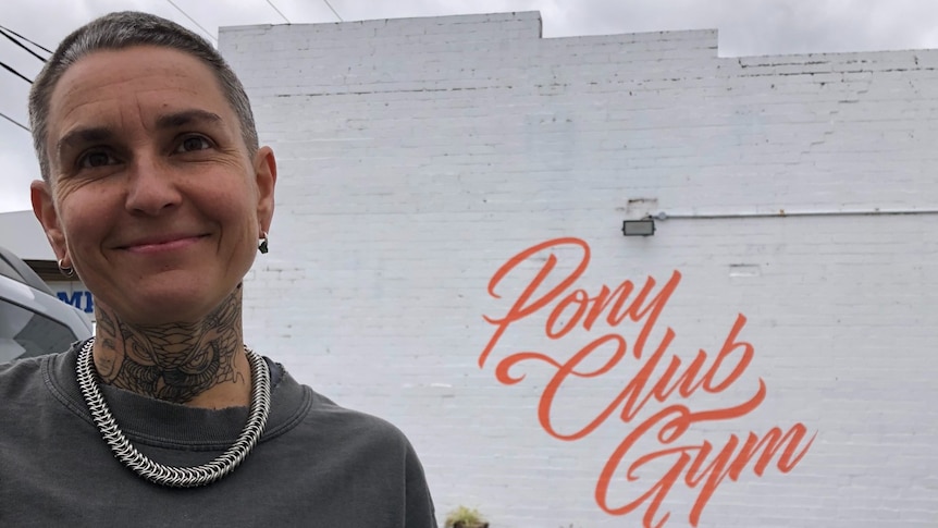 Ella Mason stands infront of the sign Pony Club Gym painted on a white, brick wall