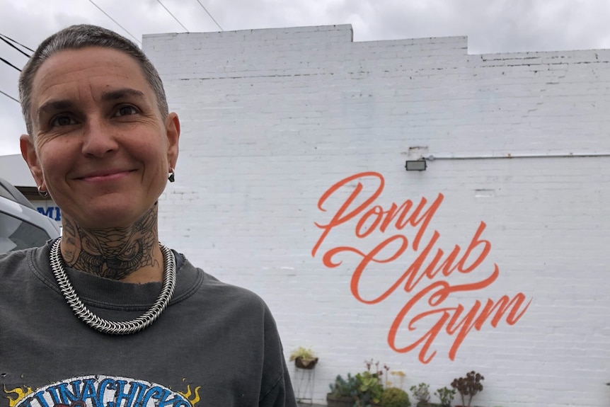 Ella Mason stands infront of the sign Pony Club Gym painted on a white, brick wall