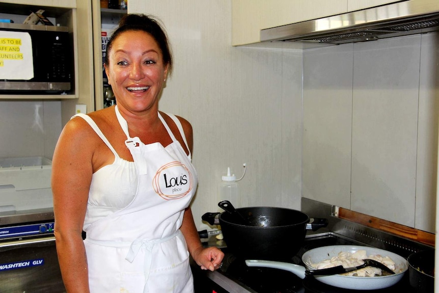A woman in an apron smiles while standing next to a stove