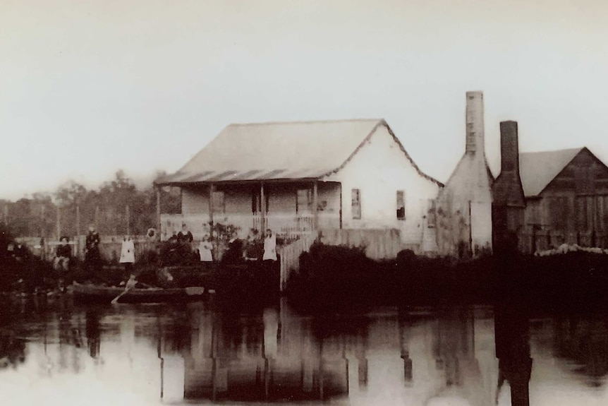 A black and white photo of an old wooden house on the banks of a river.