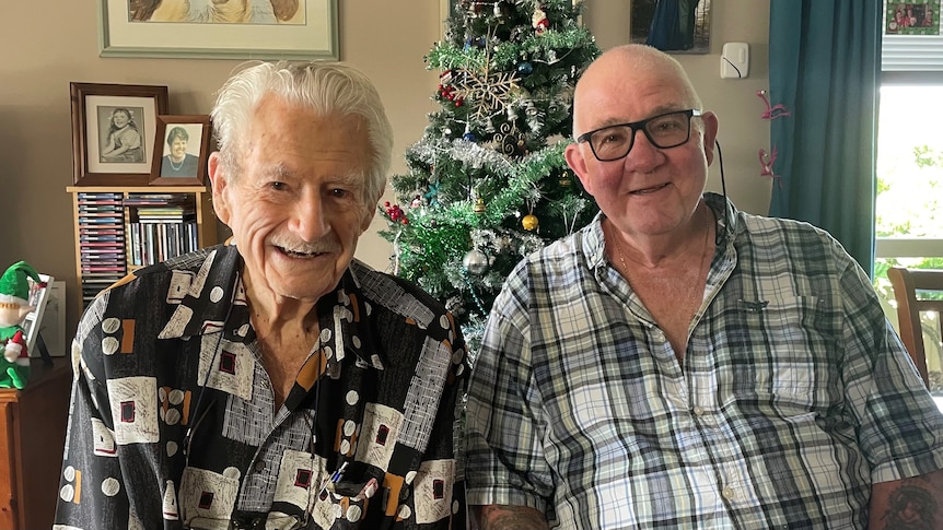 An older man and his son sit next to each other in front of a Christmas tree.