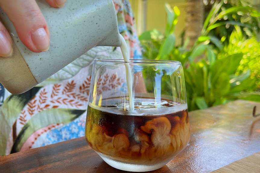 Hand holding milk jug pours milk into glass coffee cup